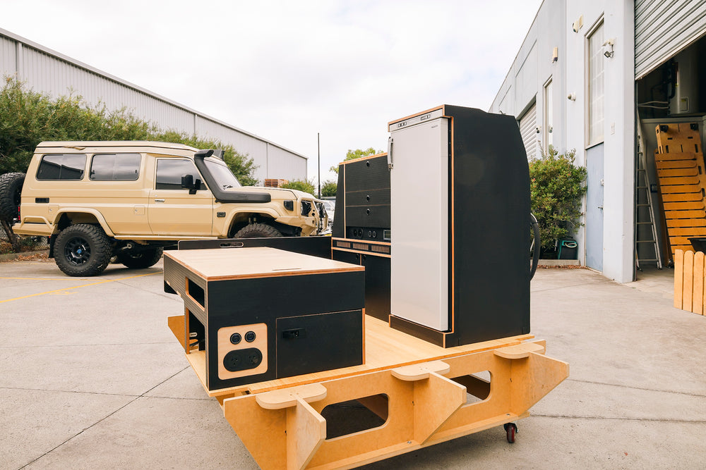 Black hex and ply Troopy cabinetry with an upright fridge on a workshop stand, a LandCruiser parked behind