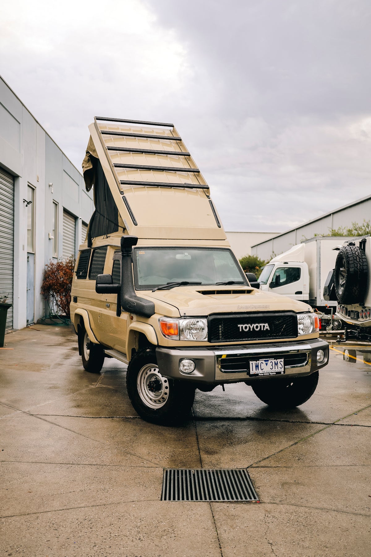 Sand-coloured late-model Troop Carrier with its pop-top raised, parked outside a workshop