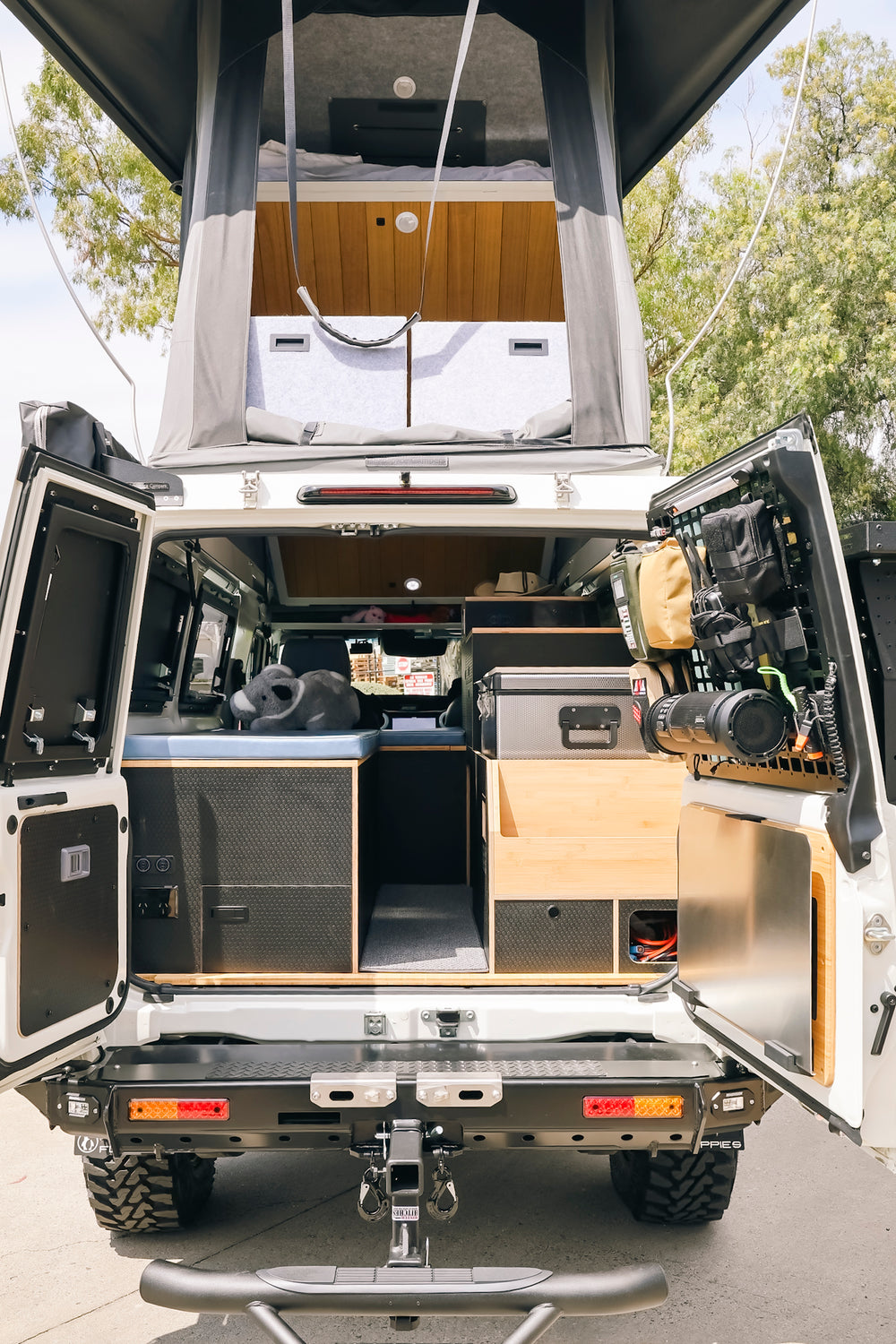 White Troopy from the rear with the roof up and barn doors open, showing black and bamboo cabinetry and a fridge